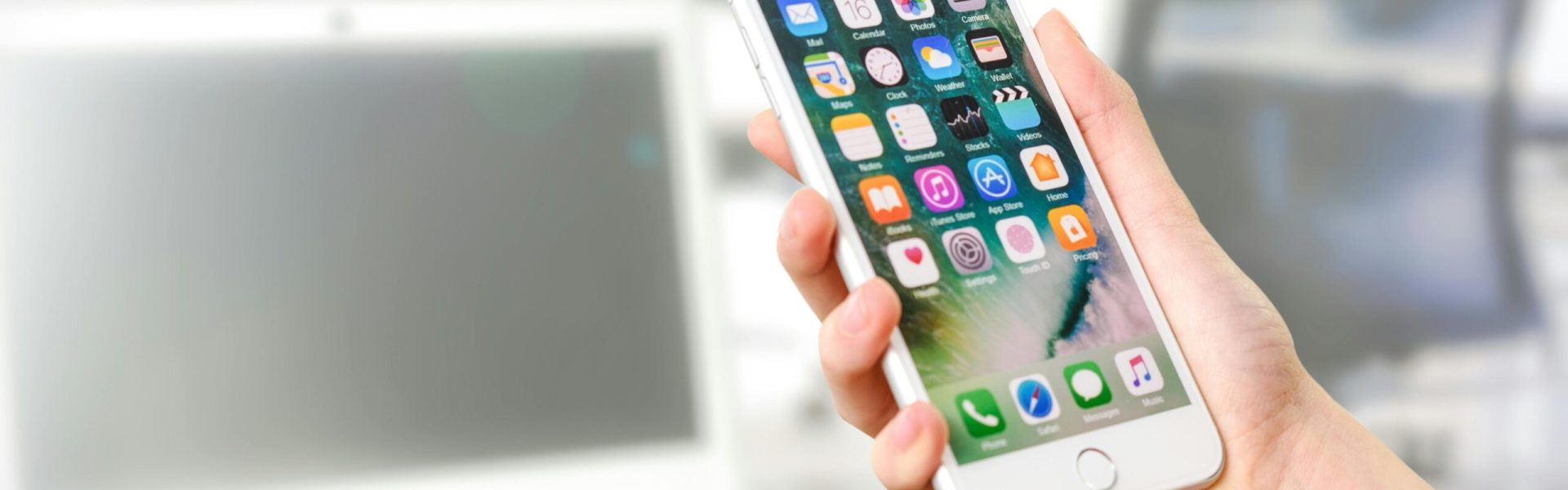 Close-up view of a smartphone displaying apps, held by a hand, with a blurred laptop in the background.